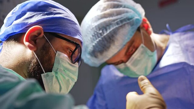 Bearded male doctor wearing glasses, cap and mask working at surgery. Side view portrait of a surgeon focused on operation.