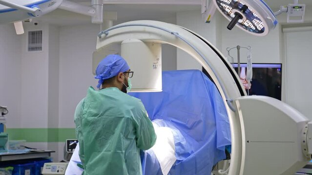 Rear view of a bearded doctor standing at C-Arm machine set above the patient. Another surgeon comes up to an operational table.