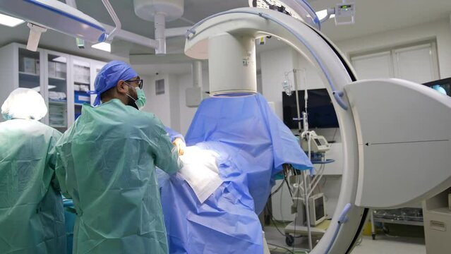 Surgeon wearing glasses and mask doing an x-ray shot of a patient on the operational table. C-Arm machine is removed from the operational field.