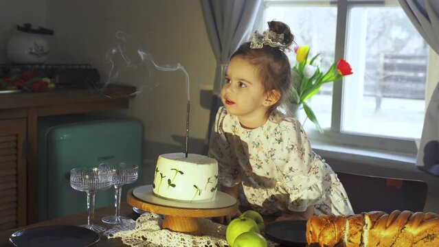A young girl in a party hat blowing out colorful candles on a birthday cake surrounded by excited friends and family.