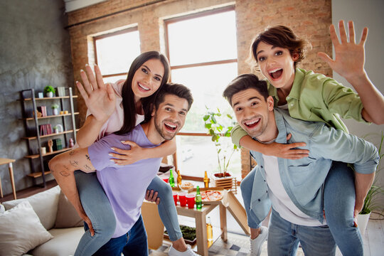 Photo of overjoyed cheerful friends hold piggyback have good mood arms waving hi free time apartment inside