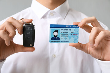 Young man holding driver license and car key on grey background, closeup