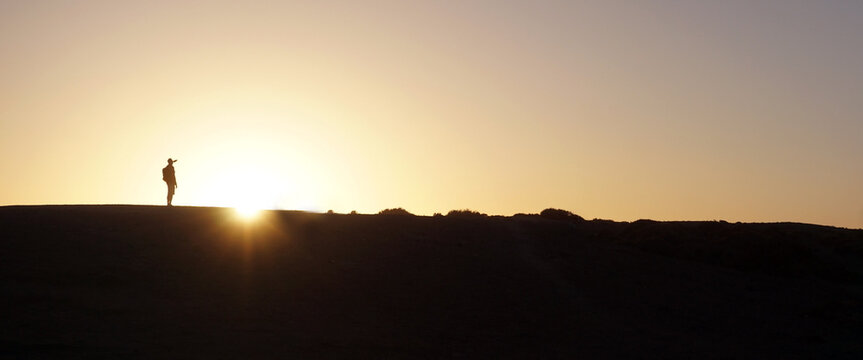 Solitary figure stands on a dune at sunrise, backlit against a warm golden sky. A peaceful, minimalist landscape evoking solitude, reflection, freedom.