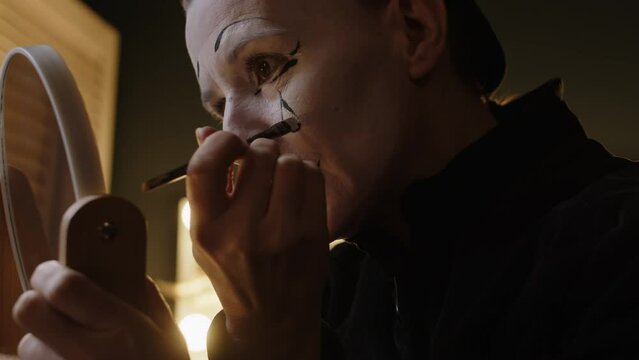 Chest up of mature female mime artist looking in mirror and painting tear on her face while doing stage mimic makeup in backstage room
