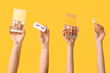 Female hands with eyeshadows palettes, lip gloss and fake lashes on yellow background