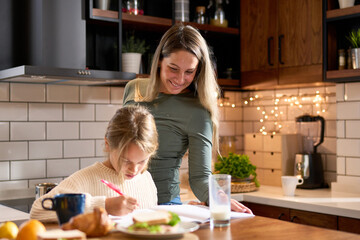 A little girl, pencil in hand, works on homework as her mother assists, blending breakfast and...