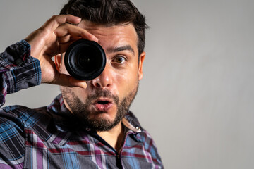 Hombre mirando a través de lente de cámara fotográfica haciendo un gesto de sorpresa y asombro - obrazy, fototapety, plakaty