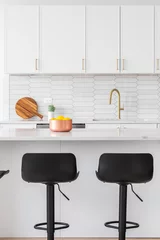 Fotobehang A kitchen detail with white cabinets, a picket tiled backsplash, gold faucet and hardware, and bar stools sitting at the marble island. © Joe Hendrickson