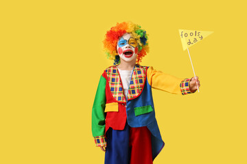Funny little boy in clown costume with paper flag on yellow background. April Fools' Day celebration