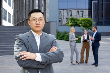 A poised businessman stands with arms crossed as his team discusses in the urban setting, embodying...