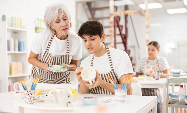 Interested elderly woman assisting young adult son working on painting ceramic mugs in family workshop. Concept of intergenerational exchange of pottery craftsmanshi