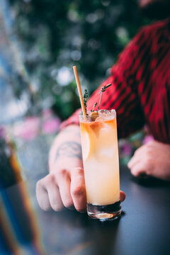 A man in a red shirt holds a classic gin cocktail garnished with orange fruit, against a background of summer flowers. Bright light adds to the atmosphere of celebration and enjoyment.