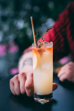 A man in a red shirt holds a classic gin cocktail garnished with orange fruit, against a background of summer flowers. Bright light adds to the atmosphere of celebration and enjoyment.