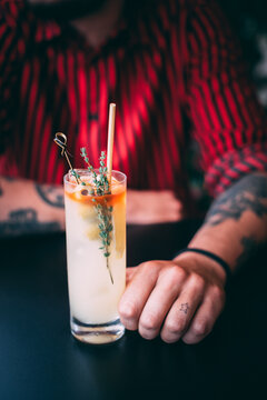 A man in a red shirt holds a classic gin cocktail garnished with orange fruit, against a background of summer flowers. Bright light adds to the atmosphere of celebration and enjoyment.