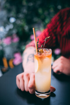 A man in a red shirt holds a classic gin cocktail garnished with orange fruit, against a background of summer flowers. Bright light adds to the atmosphere of celebration and enjoyment.