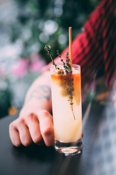 A man in a red shirt holds a classic gin cocktail garnished with orange fruit, against a background of summer flowers. Bright light adds to the atmosphere of celebration and enjoyment.