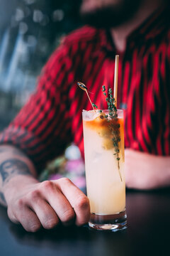 A man in a red shirt holds a classic gin cocktail garnished with orange fruit, against a background of summer flowers. Bright light adds to the atmosphere of celebration and enjoyment.