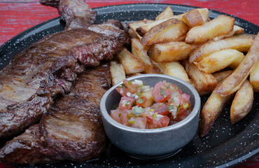 Closeup view of skirt steaks with french fries and criolla sauce, in a black dish.