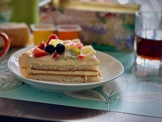 Delicious Slice of Fruit-Topped Sponge Cake on a Floral Plate with Refreshing Beverage in the Background