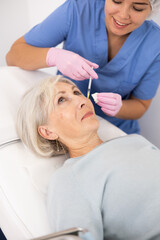 Obraz na płótnie Canvas Worried old woman lying on clinical chair in aesthetic cabinet during facial mesotherapy procedure by injection