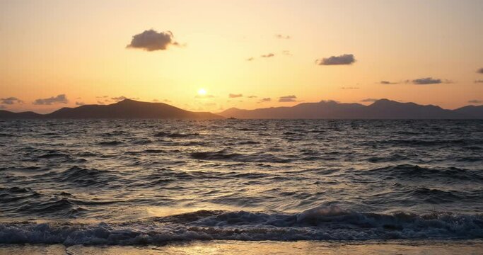 This captivating video captures a perfect moment in nature. While the calm waves of the sea sparkle under the slowly setting sun, the silhouette of the mountains blending into the shadows can be seen 