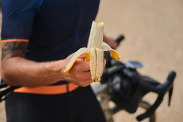 Foto op Plexiglas Man cyclist holding a banana with a background of a blue cycling jersey. Healthy nutrition of a cyclist. Healthy snack for a cyclist during training. © Ketrin