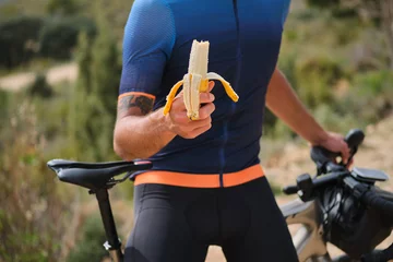 Foto op Plexiglas Man cyclist holding a banana with a background of a blue cycling jersey. Healthy nutrition of a cyclist. Healthy snack for a cyclist during training. © Ketrin