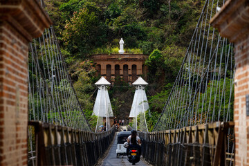 Motorcycles cross the West suspension Bridge in Santa Fe de Antioquia, Colombia. Focus on the statue of the virgin on the eastern side