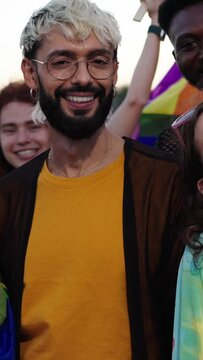 Vertical group of people are standing next to each other holding rainbow flags, smiling with happy facial expressions. They are wearing purple shirts, hats, and beards, ready to having fun photograph