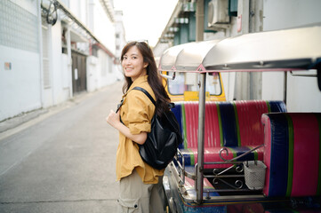 Young Asian woman backpack traveler standing a side of Tuk Tuk taxi on summer vacations at Bangkok, Thailand. Journey trip lifestyle, Asia summer tourism concept.