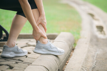 Sprained ankle problem. woman jogger. 30s asian female wearing sportswear holding her ankle with pain after running exercise in public park.