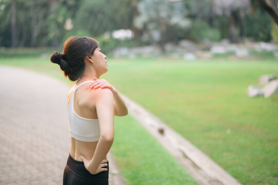 Shoulder pain problem. woman jogger. 30s asian female wearing white sportswear holding her shoulder with pain after running exercise in public park.