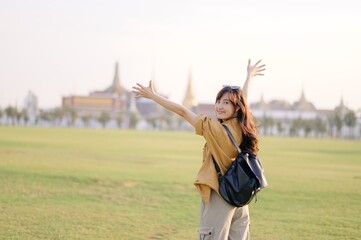 A Traveler Asian woman in 30s, bathed in golden glow of a Bangkok sunset, laughs with carefree joy, her arms outstretched as if to embrace freedom of the moment. Backpack strapped on.