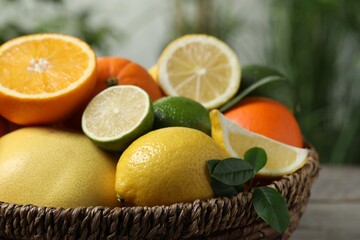 Different fresh citrus fruits and leaves in wicker basket against blurred background, closeup