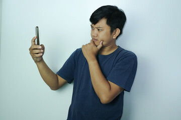 Young Asian man poses suspiciously while looking at a smartphone