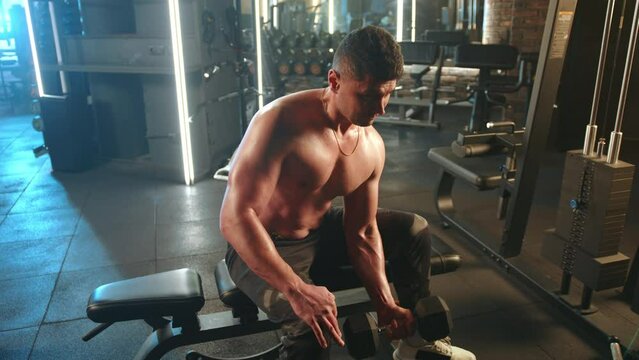 Focused young man with bare chest building muscle lifting a dumbbell in a sunlit modern gym setting. His dedication to fitness and strength training is evident. Camera 8K RAW. 