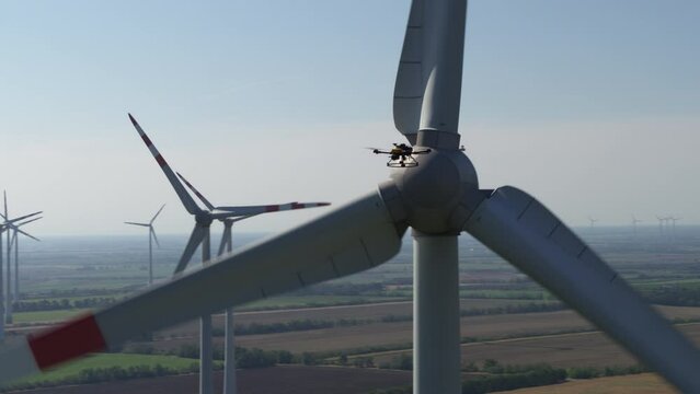 A drone in the air surveying a working wind turbine to the backdrop of a wind farm on flat farm land