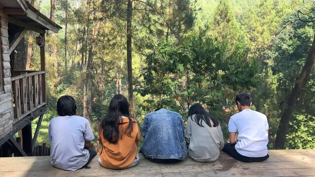 Rear View Of Young Friends Sitting at Wooden House Enjoying Grenery Scenic at The Forest During Holiday. Raising Hands Together Feel Freedom. 