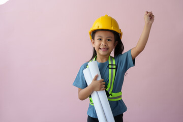 A cute girl wearing a yellow construction hat or safety helmet standing on a pink background holding a blueprint. Construction equipment, design, future career concepts, engineers, architects.