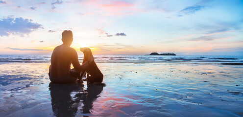 dog and human sitting together on the beach at sunset, friendship, silhouette of man with his dog, banner background with copyspace