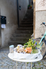 A bicycle with a flat tire is parked next to a table topped with a flowerpot, homemade jams and canned goods for sale near street stairs. The colorful flowers contrast with the worn road surface