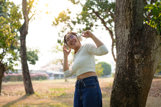 A young Asian woman wears glasses and headphones, using her mobile phone to listen to music, dance, and chat online happily, enjoying herself while relaxing on the grass in the garden. Holiday concept