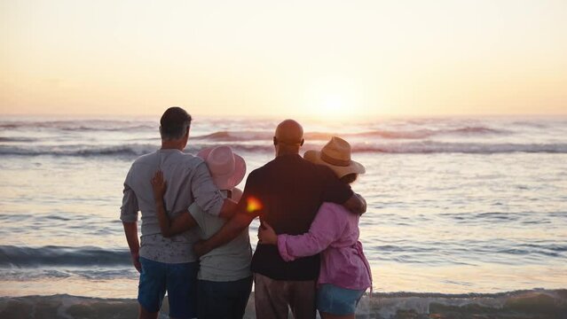 Camera pushes in on rear view of mature couple with friends standing on beach shoreline looking out to sea at sunset - shot in slow motion