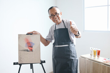 Senior Asian man wear glasses drawing canvas at home, smiling happy and showing thumb up doing...