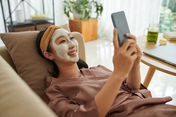 Happy woman with clay mask on face relaxing on couch and texting friends