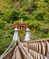 Naklejka premium Iconic Puente de Occidente in Santa Fe de Antioquia, Colombia, with a statue overlooking the historic structure amidst lush greenery