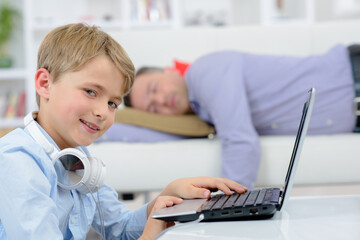Child on laptop giving cheeky smile as father sleeps