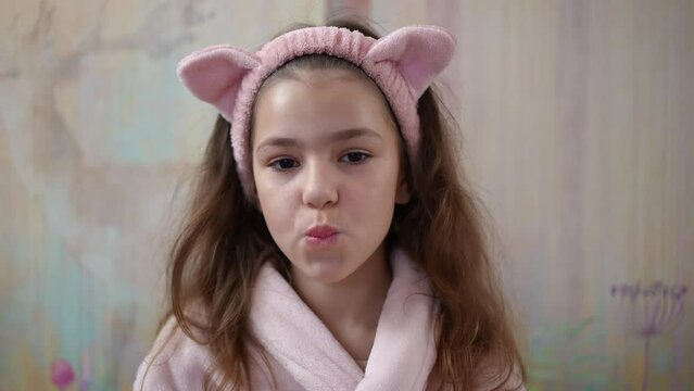 Slow motion. A little girl, in a bathrobe with a pink cosmetic hair band on her head, looks at the camera, purses her lips, makes a kiss and smiles