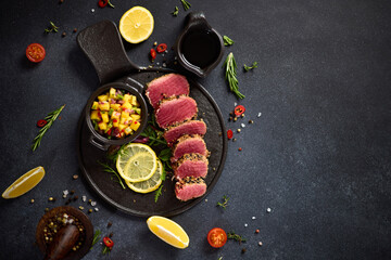 Sliced Organic grilled Tuna fillet covered with sesame seeds and mango salsa on black ceramic serving board