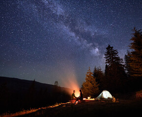 Night-time camping retreat in mountains, under celestial canopy. Man and woman trekkers relax by...